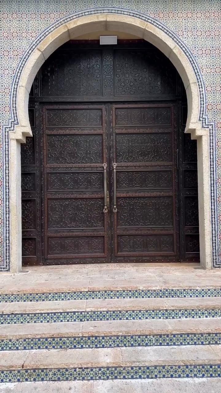 Carved wooden double door in a Moroccan-style horseshoe arch with zellige tiles, exhibition venue