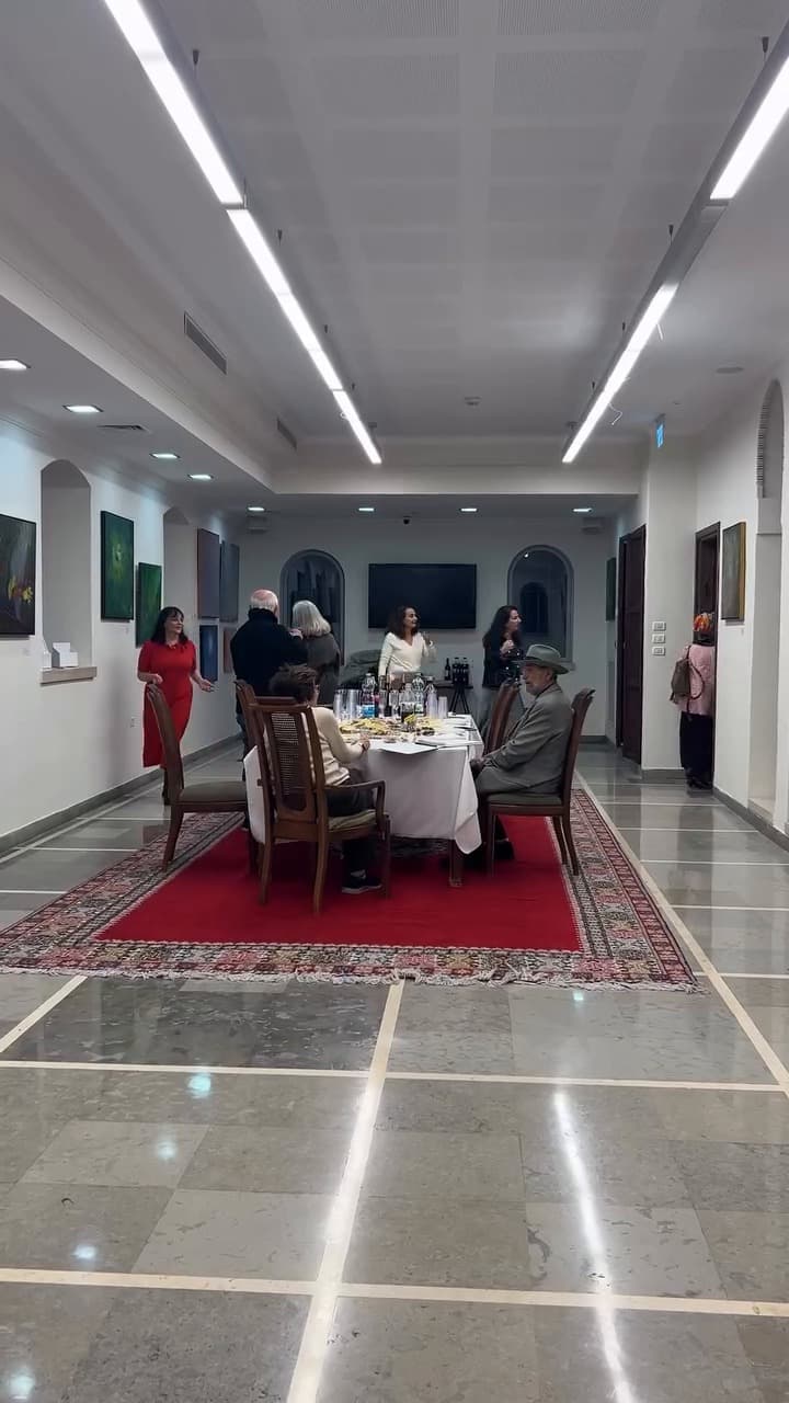 Exhibition hall with paintings on the walls and guests around a table on a red rug, Échappées belles